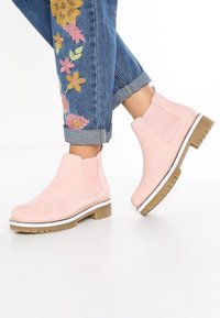 Różowe zamszowe botki na kostkę z elastycznymi panelami po bokach i białą podeszwą środkową, z charakterystyczną, ruggedową brązową podeszwą. Noszone z podwiniętymi dżinsami.