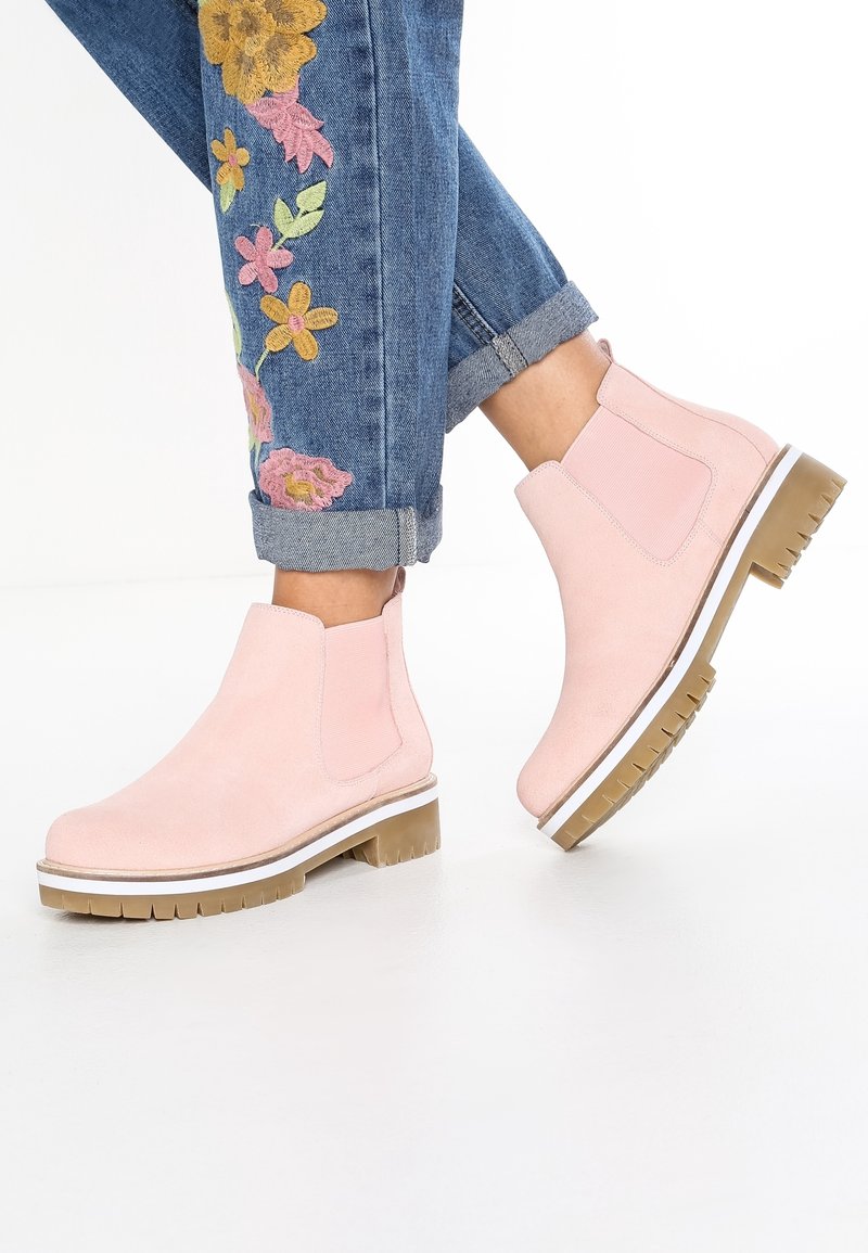 Różowe zamszowe botki na kostkę z elastycznymi panelami po bokach i białą podeszwą środkową, z charakterystyczną, ruggedową brązową podeszwą. Noszone z podwiniętymi dżinsami.