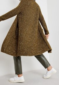 Langer, olivgrüner Strickcardigan mit gerippter Textur und subtiler Glanz, getragen über dunklen Lederhosen und weißen Sneakern.