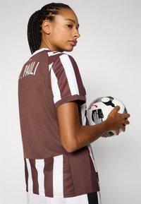 Brown and white striped sports jersey with short sleeves, featuring the name "St. Pauli" on the back. Model holds a black and white soccer ball.