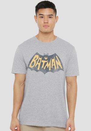 Young man facing forward wearing a grey t-shirt with a yellow and black Batman logo and beige pants against a plain background.