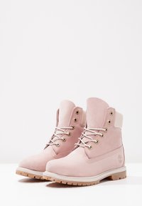 Une paire de bottines lacées rose clair avec des cols rembourrés et des semelles robustes couleur tan, présentées sur une surface blanche avec un fond uni.