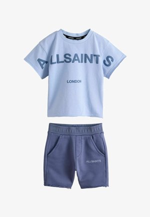 Młody chłopiec stojący z rękami w kieszeniach, ubrany w jasnoniebieski t-shirt AllSaints London, ciemne szorty, białe skarpetki i białe trampki.