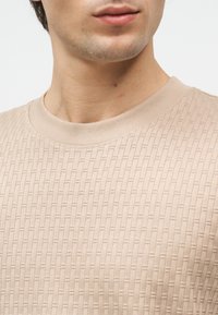 Κοντινό πλάνο ατόμου που φοράει μπεζ πλεκτό πουλόβερ με στρογγυλή λαιμόκοψη, εμφανίζοντας τον λαιμό, το κάτω μέρος του προσώπου και μέρος του άνω στήθους.