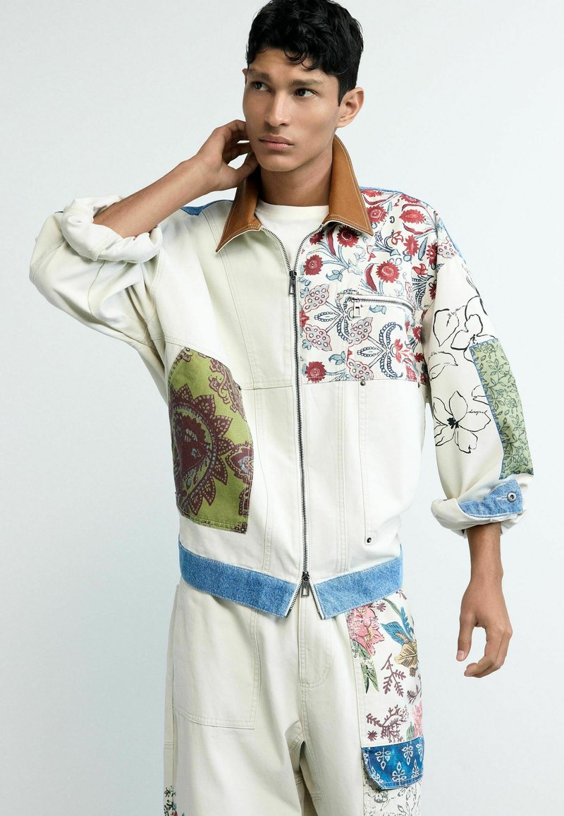 Noor mees, kes kannab kreemikat jopet ja pükse mitmevärviliste lille- ja plaastrimustritega, üks käsi toetub kaela taha.