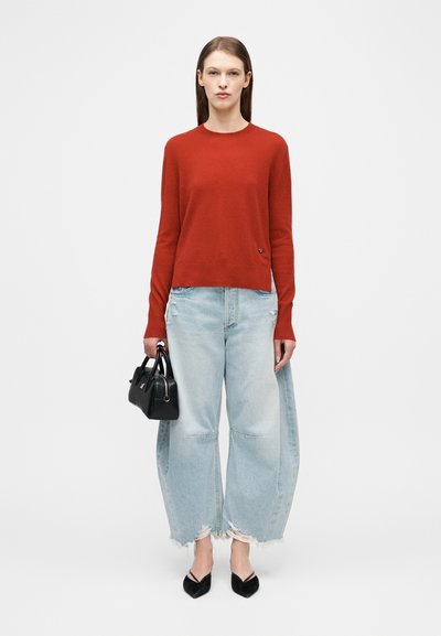 Γυναίκα που φοράει κόκκινο πουλόβερ και φαρδιά τζιν παντελόνια σε ανοιχτό μπλε χρώμα με σκισίματα, κρατώντας μια μαύρη τσάντα, στέκεται μπροστά σε απλό φόντο.
