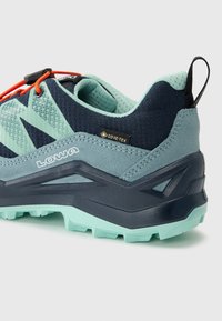 Atletska cipela u tirkiznoj i tamnoplavoj boji s teksturiranim gornjim dijelom, GORE-TEX oznakom, ojačanim petnim dijelom i kontrastnim narančastim vezicama.