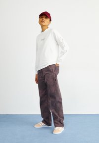 Camisa blanca de manga larga con detalles gráficos, combinada con pantalones cargo marr�ón oscuro y zapatillas beige claras, todo sobre un fondo liso.