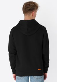 Jonge man met kort, krullend haar, gekleed in een eenvoudige zwarte hoodie en lichtblauwe jeans, met zijn rug naar de camera gericht tegen een witte achtergrond.