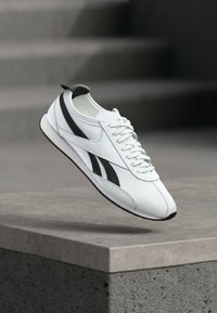 Tenis alb cu dungi negre și sireturi, suspendat deasupra unui margine de beton, cu trepte întunecate neclare în fundal.