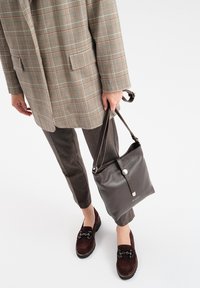 Brązowa skórzana torba w kształcie wiadra z teksturowanym wykończeniem, srebrnymi detalami i regulowanym paskiem, w komplecie z marynarką w kratę i brązowymi spodniami.
