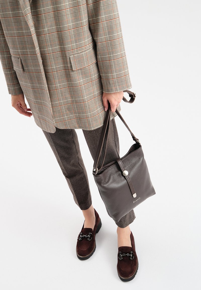 Brązowa skórzana torba w kształcie wiadra z teksturowanym wykończeniem, srebrnymi detalami i regulowanym paskiem, w komplecie z marynarką w kratę i brązowymi spodniami.