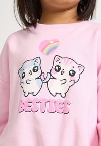 Felpa rosa chiaro con una grafica di due gatti cartoni animati, uno blu e uno bianco, che si tengono per la coda. Il testo recita "BESTIES" in lettere grasse e arrotondate.