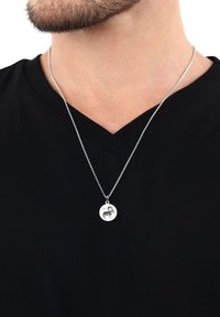 Uomo che indossa una collana d'argento con un pendente rotondo raffigurante una capra, sopra una maglietta nera a scollo a V.