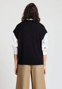 Schwarzes, ärmelloses Stricktop mit Rundhalsausschnitt. Wird über einem weißen Langarmshirt getragen und mit beigen, weit geschnittenen Hosen kombiniert.