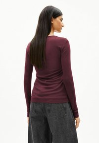 Bordeauxrotes Langarmshirt mit figurbetontem Schnitt, aus weichem Material. Die Rückseite zeigt eine nahtlose Optik und einen abgerundeten Ausschnitt, kombiniert mit dunklen Jeans.