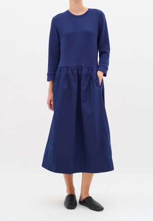 Kobieta ubrana w granatową sukienkę midi z długim rękawem i marszczonym pasem, stojąca na tle jednolitego białego tła, w czarnych wsuwanych butach.