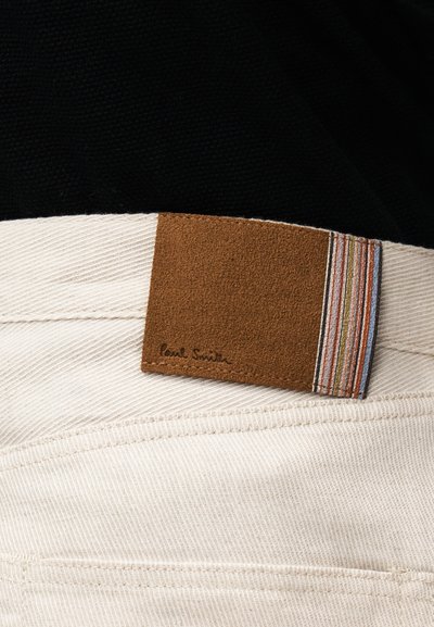 Gros plan sur un pantalon en velours côtelé beige avec un patch en cuir marron et un détail en tissu rayé multicolore à la ceinture.