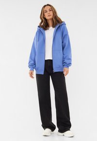Felpa blu con zip e coulisse, indossata su una maglietta bianca, abbinata a pantaloni neri a gamba larga e sneakers bianche. Tessuto di texture liscia.
