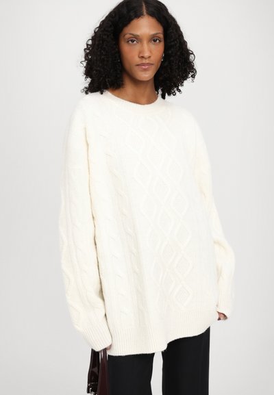 Pull en maille blanche à côtes avec une coupe ample, comportant des motifs en losange et des poignets côtelés. Associé à un pantalon foncé et un sac bourgogne.