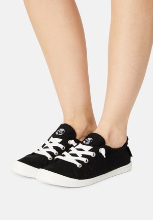 Melni auduma sneakers ar baltām šņorēm, gumijas zoli un diviem melniem apaļiem logo. Dizains ietver teksturētu virsmu un redzamām šuvēm.