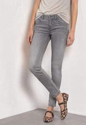 Femme portant un jean gris moulant, un haut beige et des chaussures Mary Jane imprimé léopard, debout contre un mur blanc texturé.