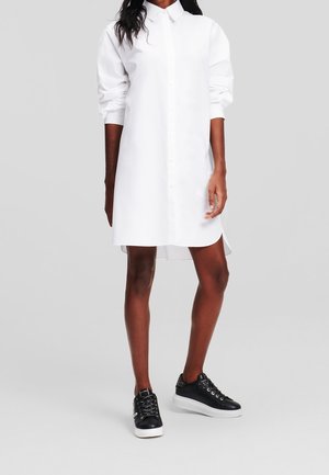 Žena oblečená v dlouhých rukávech bílých šatech připomínajících košili, spárovaných s černými nízkými teniskami s bílou podrážkou.