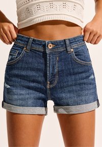 Shorts de mezclilla en azul oscuro con dobladillos enrollados. Presentan un diseño clásico de cinco bolsillos, detalles en hardware de cobre y ligeros desgastes.