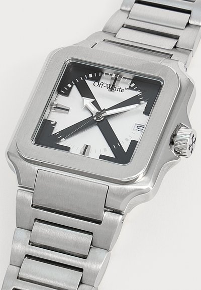 Montre-bracelet carrée en métal argenté avec un cadran noir et blanc, une aiguille des heures en forme de flèche audacieuse, une fenêtre date à trois heures et un bracelet à maillons en métal.