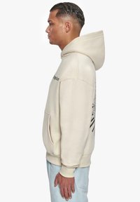 Felpa beige con grande tasca frontale, cappuccio regolabile con lacci e design grafico sul retro. Realizzata in tessuto morbido e testurizzato.