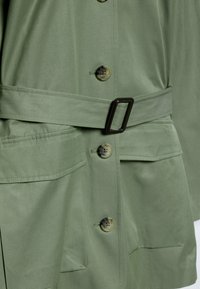 Olive green belted jacket with a smooth texture, featuring large front pockets and round buttons. Belt has a rectangular buckle.