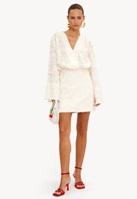 Witte textuur mini-jurk met lange mouwen, diepe V-hals en franjes aan de onderkant. Gecombineerd met een kleine witte tas met rode kersen en rode hakken.