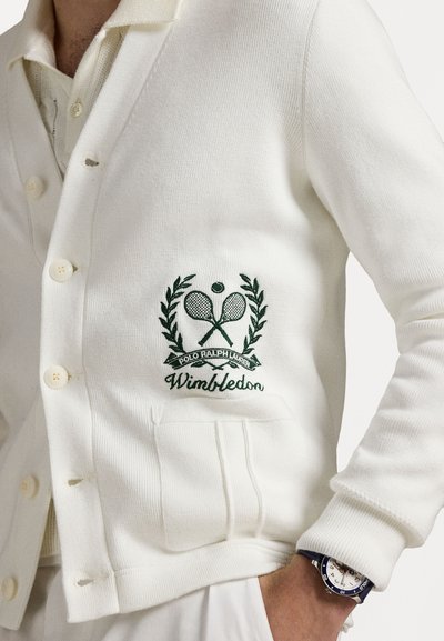 Gilet blanc à texture côtelée, avec logo Wimbledon brodé vert et deux poches avant. Les boutons sont blancs et ronds.