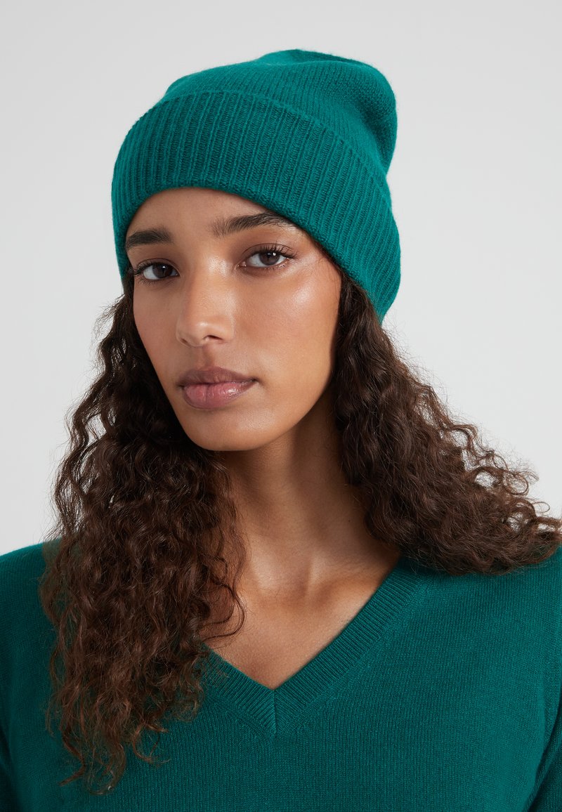 Giovane donna con capelli ricci che indossa un berretto di maglia verde teal e un maglione a V coordinato, guardando diritto davanti a sé con un'espressione neutra.
