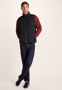 Black padded vest over a maroon long-sleeve shirt; paired with dark denim jeans and black sneakers featuring white stripes.