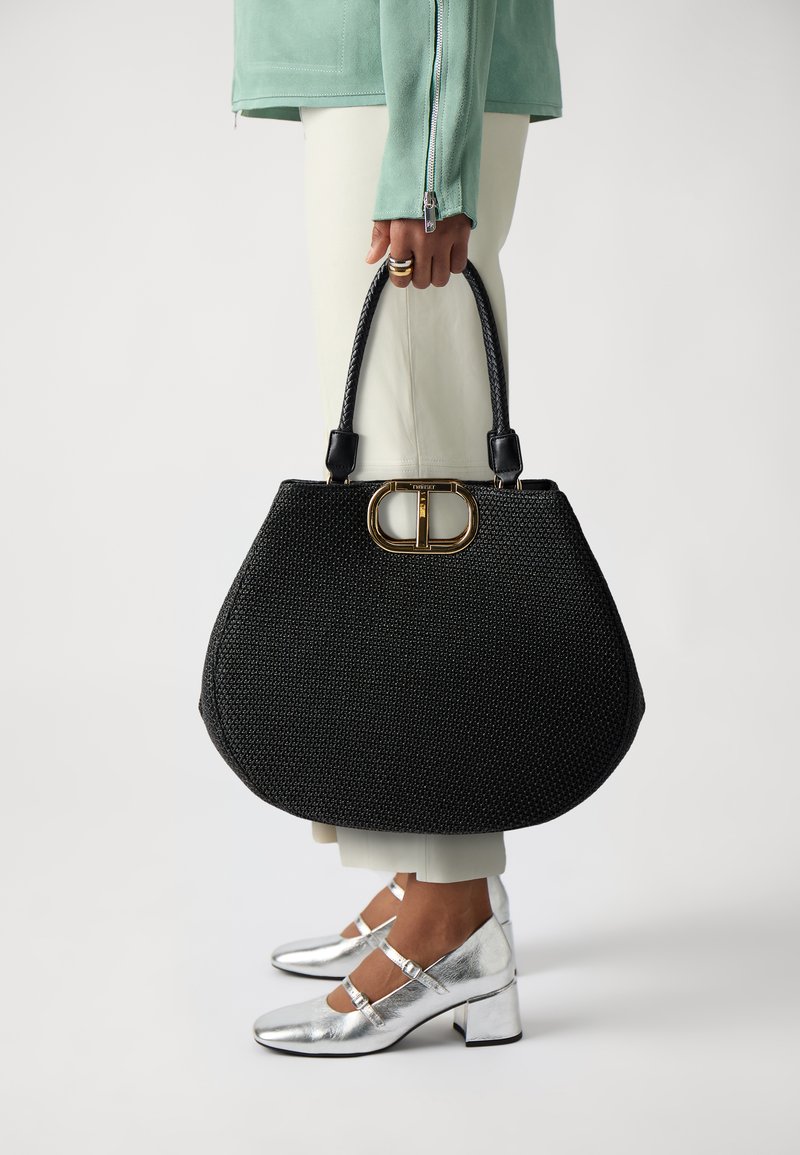 Borsa nera textrurizzata con manici corti doppi e una chiusura con accento dorato, abbinata a scarpe con tacco a blocco argentato e pantaloni crema.