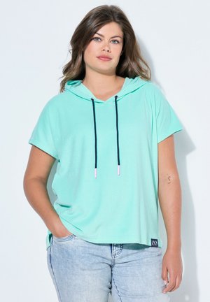 Kobieta o jasnobrązowych włosach, ubrana w miętowo zieloną bluzę z kapturem i krótkim rękawem z czarnymi troczkami oraz jasnoniebieskie jeansy, stoi z jedną ręką w kieszeni.