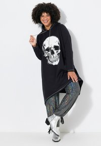 Zwarte hoodie jurk met een grote witte schedelprint, gecombineerd met glanzende, meerkleurige sequined skinny broeken en zilveren enkellaarzen.