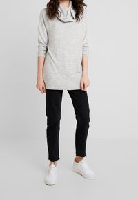 Hellgrauer, langärmeliger Pullover mit einem Schalkragen. Kombiniert mit schwarzen, schmal geschnittenen Jeans und weißen Sneakers. Der Stoff wirkt weich und strukturiert.