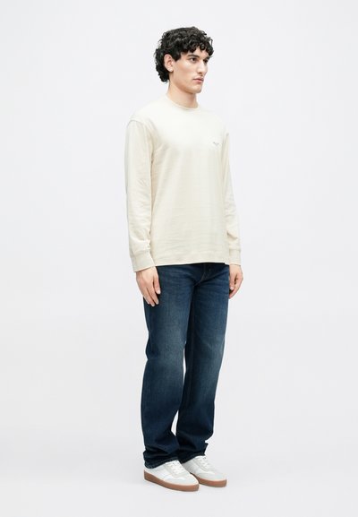 Gaiši dzeltena kokvilnas t-krekls ar garām piedurknēm un apaļu kakla izgriezumu, kopā ar tumši zilām džinsa biksēm un baltām sporta kurpēm ar brūnām zolēm.