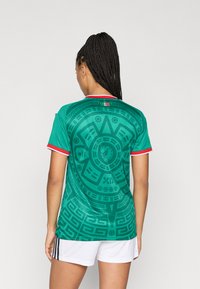 Camisa de futebol verde com padrões intrincados em preto, detalhe vermelho na gola e mangas curtas. Usada com calções brancos.