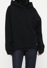 Sudadera negra con capucha, de ajuste holgado, mangas largas y tejido texturizado. Detalles notables incluyen un dobladillo curvo y una sutil marca lateral.