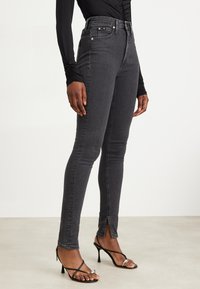 High-waisted black jeans in stretch denim with a slim fit, featuring front pockets and a slight slit at the ankle. Styled with black heels.
