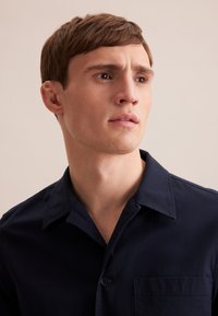 Camicia blu navy con bottone, maniche corte, tasca sul petto e una texture morbida e liscia. Collo classico e linee di design pulite.