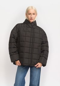Geacă pufă neagră cu guler înalt, design matlasat, croială supradimensionată și mâneci late. Dispune de buzunare laterale și detalii minime privind cusăturile.