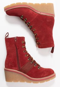 Botines tobilleros de ante rojo con un diseño de cordones y cremallera lateral. Presentan una suela de plataforma acanalada y detalles marrones en los laterales.