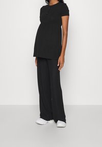 Mujer embarazada vestida con una blusa negra de manga corta y pantalones anchos, con zapatillas blancas, de pie frente a un fondo claro y liso.