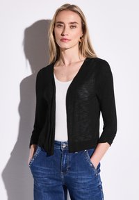 Musta, kevyt neuletakki, jossa on kolme neljäsosaa hihat, avoin etuosa, hieman teksturoitu kangas, yhdistettynä valkoiseen puseroon ja sinisiin farkkuihin.