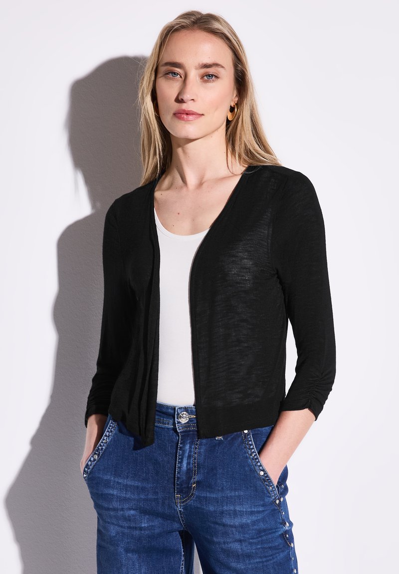 Musta, kevyt neuletakki, jossa on kolme neljäsosaa hihat, avoin etuosa, hieman teksturoitu kangas, yhdistettynä valkoiseen puseroon ja sinisiin farkkuihin.