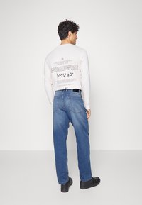 Camisa de manga longa bege clara com texto gráfico nas costas, combinada com jeans azul e sapatos pretos, vista de trás.